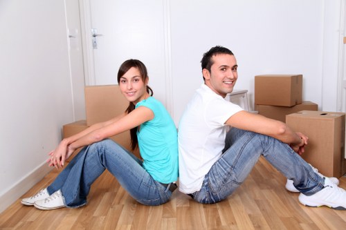 A young man and woman are seated back-to-back on a light wooden floor in a bright, uncluttered room, surrounded by neatly stacked cardboard boxes of various sizes, indicating a house moving or relocation process. The woman, on the left, is wearing a light blue t-shirt and blue jeans with white sneakers, and is smiling gently while resting her arms on her knees. The man, on the right, is dressed in a white t-shirt and blue jeans, also with white sneakers, and is smiling broadly. The background features a white wall and a closed white door, with more boxes visible behind them, suggesting an interior space before or after packing. The room is well-lit, creating a clean and organized atmosphere, typical of professional removals in the Kingston area, as part of Kingston Man with Van's home moving services.
