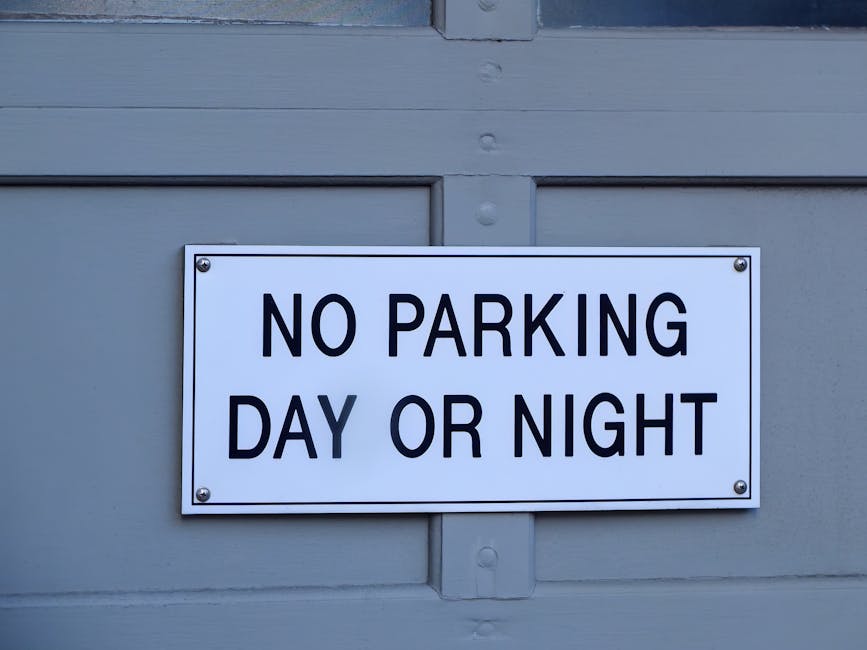 A white rectangular parking restriction sign mounted on a grey door or gate, displaying the message 'NO PARKING DAY OR NIGHT' in bold black letters. The sign is secured with four metal screws, one at 
