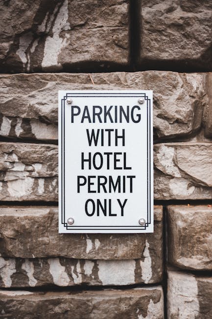A white rectangular parking restriction sign mounted on a grey door or gate, displaying the message 'NO PARKING DAY OR NIGHT' in bold black letters. The sign is secured with four metal screws, one at 